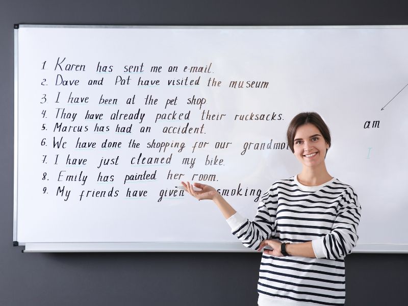 Kurs języka angielskiego od podstaw, czyli od czego zaczynamy? Na zdjęciu nauczycielka ze zdaniami angielskimi na tablicy.